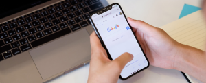 personne qui tient son téléphone et qui est sur la home page Google devant son ordinateur